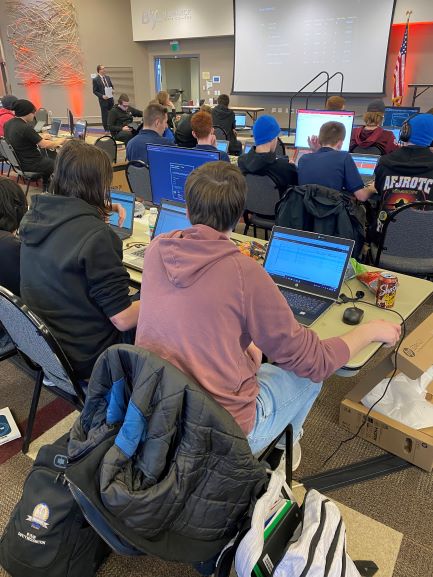 Teams of high school students sit at tables with laptops to compete in a cyber challenge.