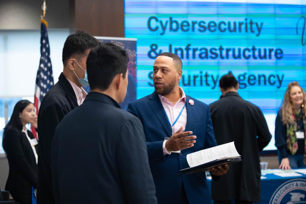 OCHCO manager talks with 2 candidates at Spring into CISA: FY24 In-Person Hiring Event, other staff and candidates are in background.