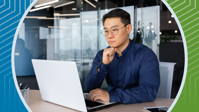 A man sits at his desk looking contemplatively towards his laptop computer